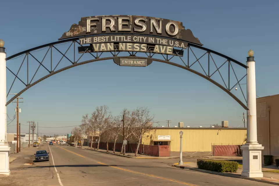 View of wide, deserted-looking street with arched sign reading "Fresno - Best little city in the U.S.A. - Van Ness Avenue - Entrance" in Fresno, California.